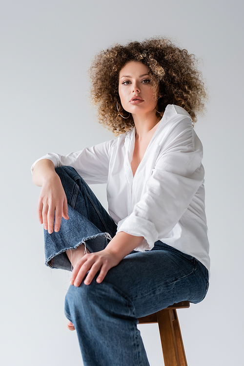 Low angle view of stylish curly woman looking at camera while sitting on chair isolated on white