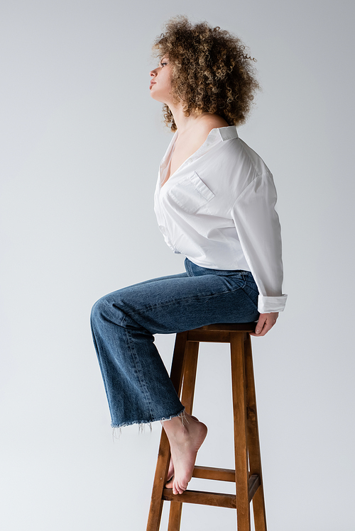 Side view of stylish barefoot model sitting on chair isolated on white