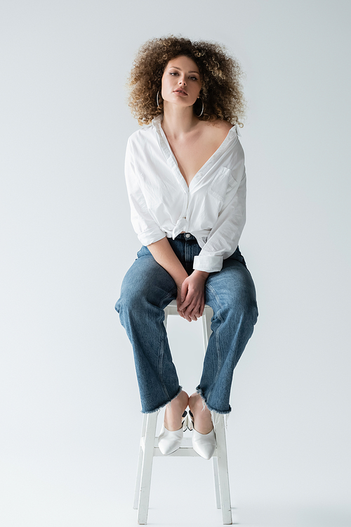 Stylish curly woman in blouse sitting on chair on white background