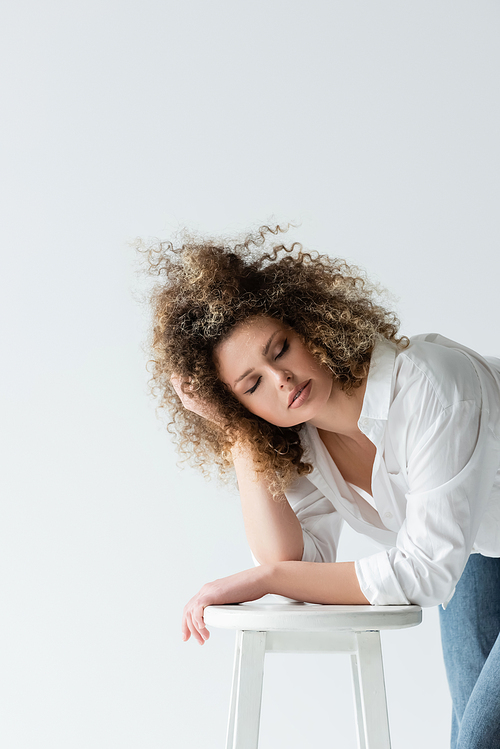 Young woman touching curly hair near chair isolated on white