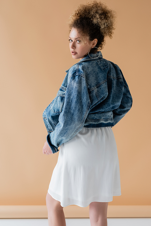 Stylish model in denim jacket looking at camera on beige background
