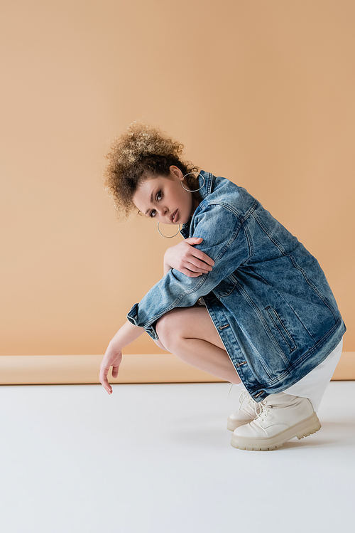 Stylish curly woman in denim jacket on beige background