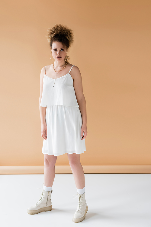 Full length of stylish woman in boots and white clothing standing on beige background