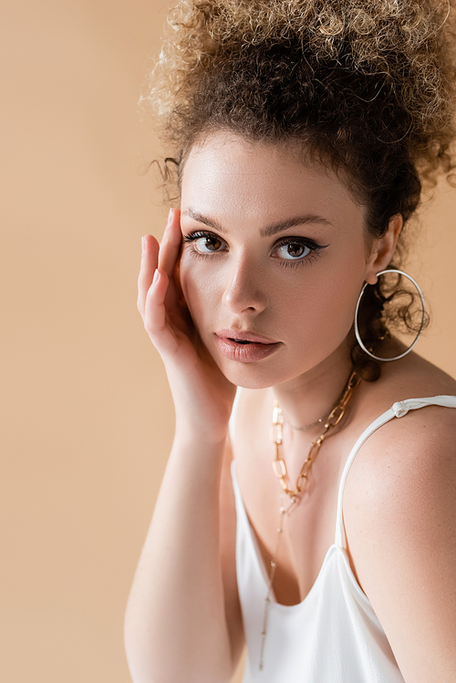 Portrait of curly model in accessories touching face isolated on beige