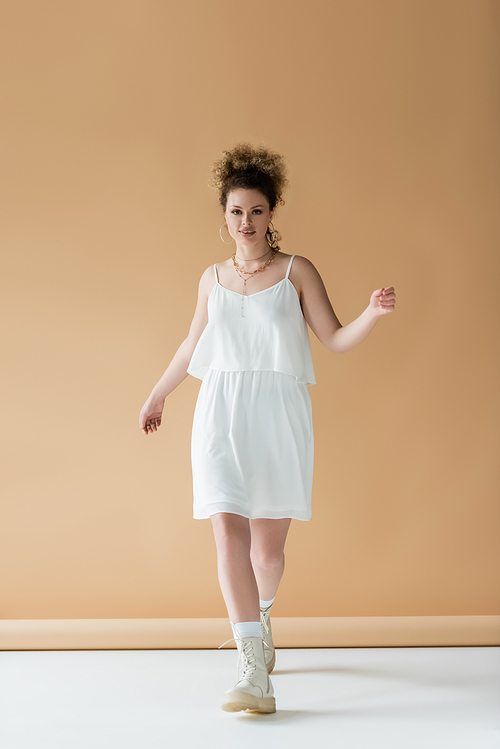 Positive woman in white clothes walking and looking at camera on beige background