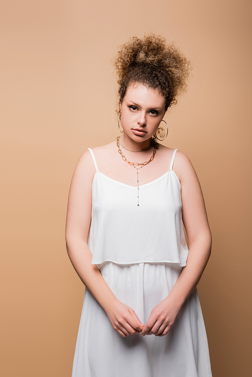 Stylish model in white clothes and necklaces looking at camera isolated on beige