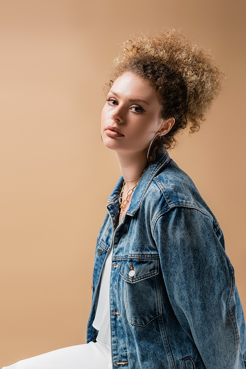 Curly model in denim jacket looking at camera isolated on beige