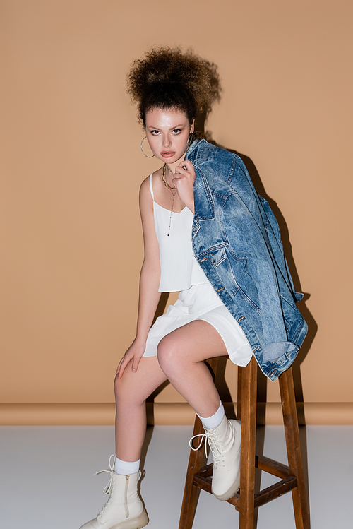 Stylish model holding denim jacket while sitting on chair on beige background