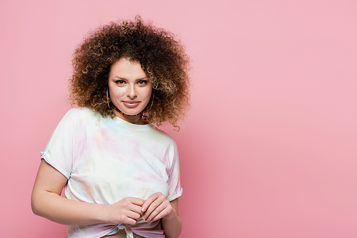 Portrait of smiling curly woman in t-shirt looking at camera isolated on pink