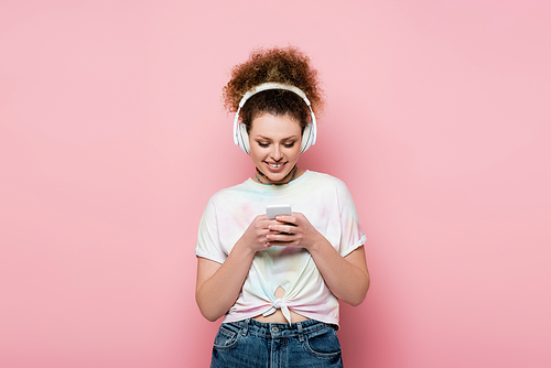 Cheerful woman in headphones using smartphone isolated on pink