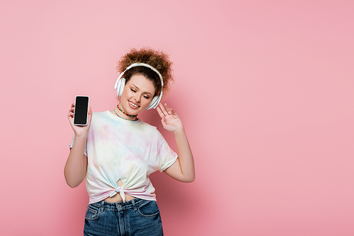 Curly woman in headphones holding cellphone on pink background