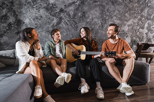 cheerful woman playing acoustic guitar near multiethnic friends sitting on couch in modern living room