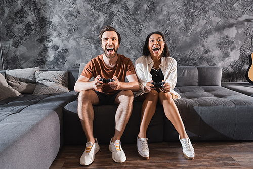 KYIV, UKRAINE - JULY 26, 2022: amazed interracial friends playing video game and sitting on grey couch