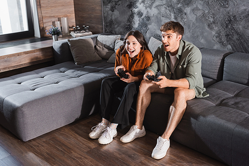 KYIV, UKRAINE - JULY 26, 2022: excited friends holding joysticks and playing video game while sitting on grey couch