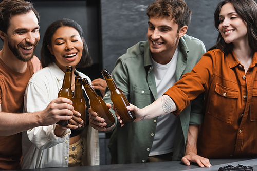 KYIV, UKRAINE - JULY 26, 2022: happy interracial friends clinking bottles of beer near joystick on kitchen worktop