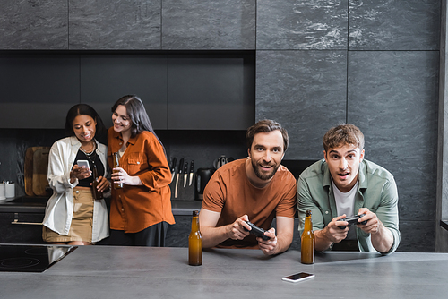 KYIV, UKRAINE - JULY 26, 2022: happy men playing video game near blurred interracial women using smartphone in kitchen