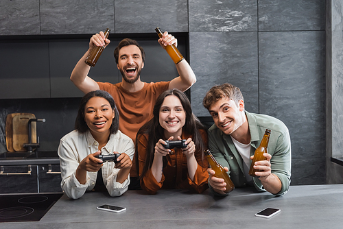 KYIV, UKRAINE - JULY 26, 2022: happy interracial women playing video game near cheerful male friends with beer in kitchen