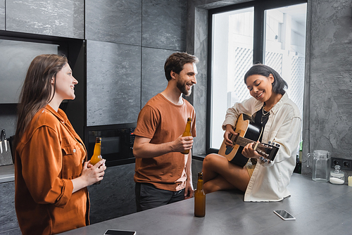 happy bi-racial woman playing acoustic guitar near friends with bottles of beer