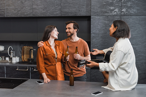 happy man hugging woman near bi-racial friend with acoustic guitar gesturing in kitchen