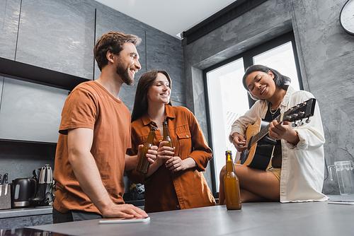 low angle view of happy bi-racial woman playing acoustic guitar near cheerful friends with bottles of beer