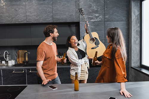 amazed bi-racial woman holding  acoustic guitar near cheerful friends with bottles of beer