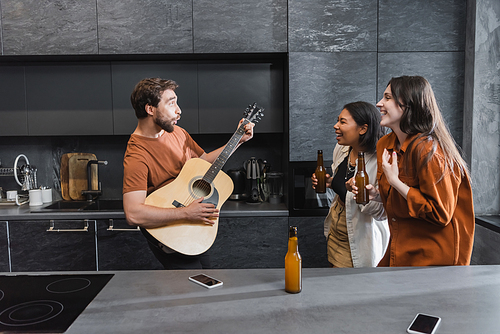bearded man playing acoustic guitar near happy interracial women with bottles of beer in kitchen