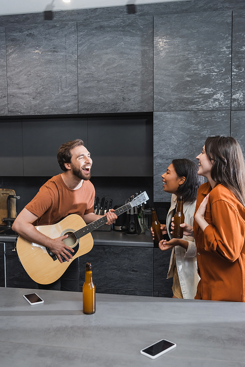 cheerful man playing acoustic guitar near happy interracial women with bottles of beer in kitchen