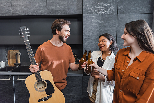 cheerful man holding acoustic guitar and clinking bottles of beer with happy interracial women in kitchen