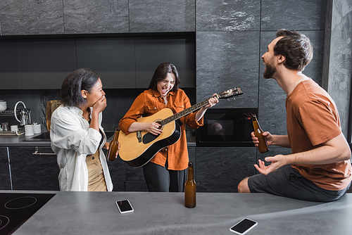 excited woman with closed eyes playing acoustic guitar near interracial friends in kitchen