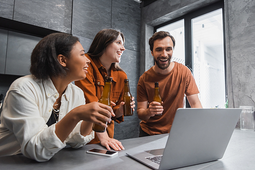 happy multiethnic friends holding bottles of beer near laptop and smartphone with blank screen on kitchen worktop