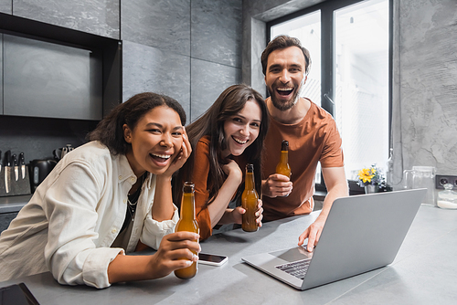 happy multiethnic friends holding bottles of beer near gadgets and laughing in kitchen