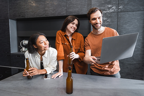 happy man holding laptop near multiethnic friends with bottles of beer in kitchen