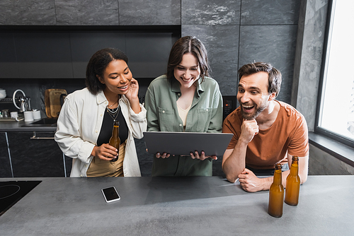 happy woman holding laptop near multiethnic friends with bottles of beer in kitchen