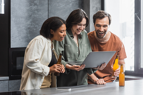 happy woman holding laptop near interracial friends with bottles of beer in kitchen