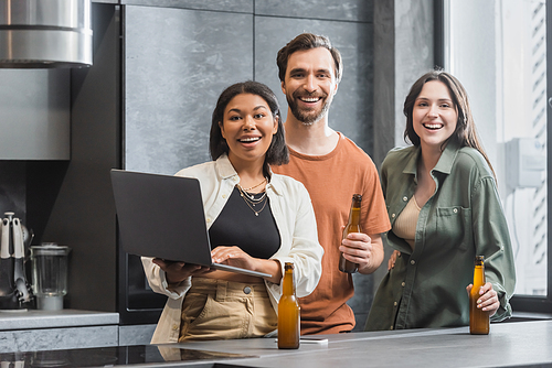 happy bi-racial woman holding laptop near friends with bottles of beer standing in kitchen