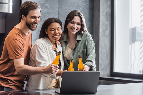 cheerful multiethnic friends holding bottles of beer and watching comedy movie on laptop