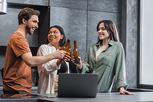 cheerful multiethnic friends holding bottles of beer and clinking near laptop in kitchen