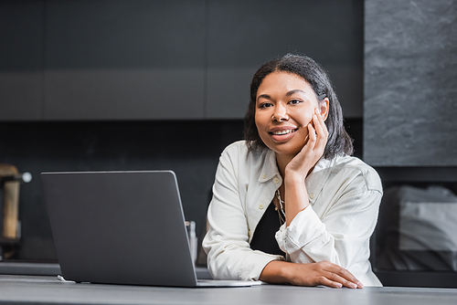 happy bi-racial woman looking at camera near laptop on kitchen worktop