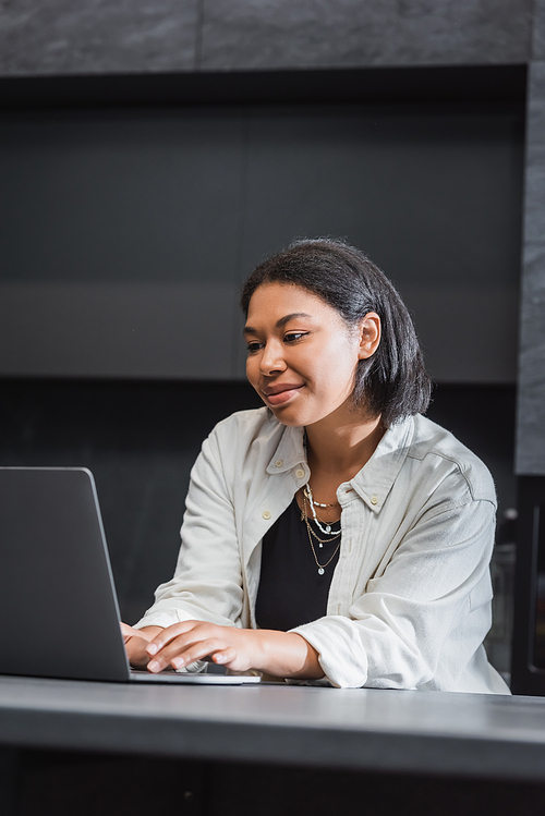 happy bi-racial woman using laptop while working remotely in kitchen