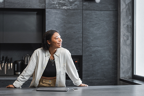 excited bi-racial woman smiling while looking away near smartphone and laptop on kitchen worktop