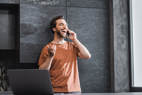 happy bearded man talking on smartphone and laughing while standing near laptop