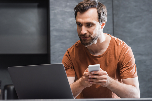 bearded freelancer holding smartphone while looking at laptop