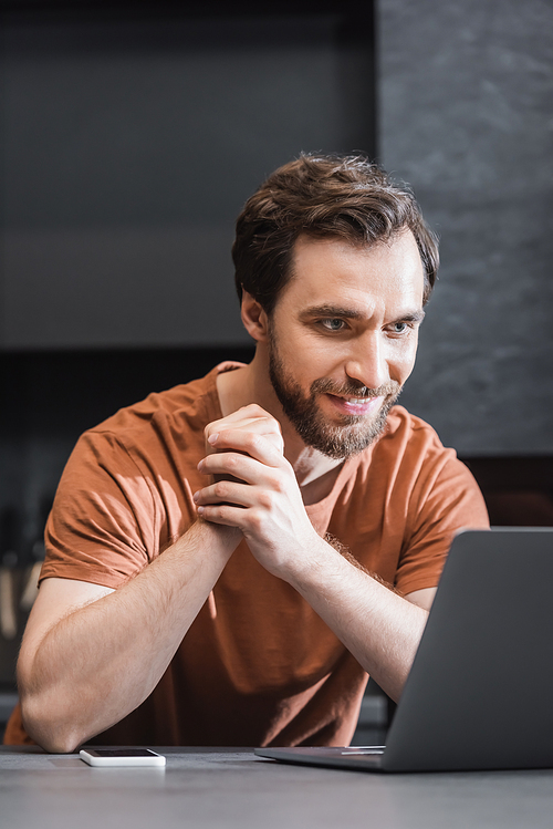 cheerful bearded freelancer looking at laptop near smartphone on kitchen worktop
