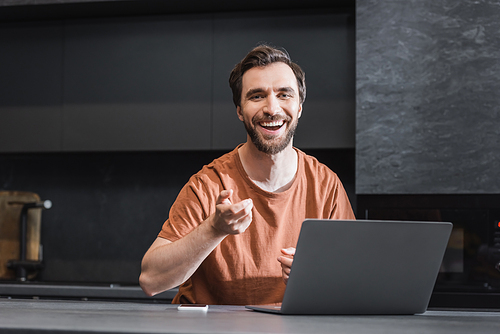 excited bearded freelancer laughing near laptop and smartphone on kitchen worktop