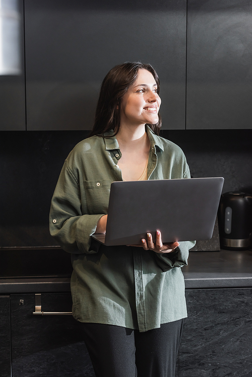 cheerful young woman in green shirt holding laptop and looking away in kitchen