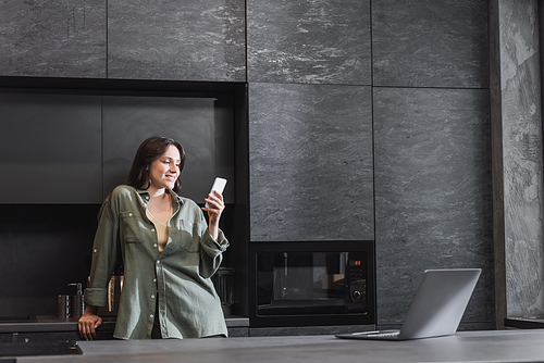 cheerful young woman in green shirt holding smartphone near laptop on kitchen worktop