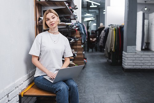 Blonde saleswoman holding laptop and looking at camera in vintage shop