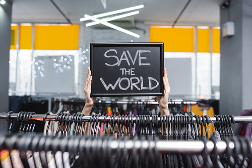 Cropped view of saleswoman holding nameplate with save the world lettering near clothes in second hand