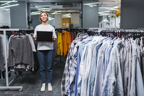Smiling saleswoman holding empty board near clothes in second hand