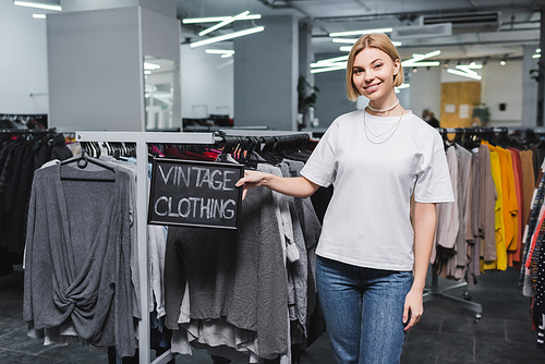 Smiling saleswoman holding board with vintage clothing lettering in retro store
